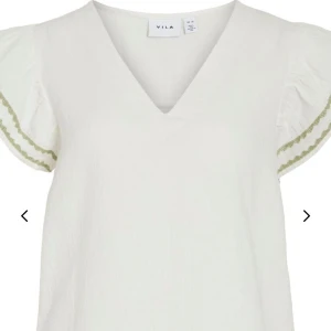 Vit topp med puffärm från Vila - Säljer en söt vit topp från Vila med puffiga kortärmar och gröna broderade detaljer på ärmarna. Toppen har en djup v-ringning och är gjord i ett lätt, strukturerat bomullstyg som känns luftigt och fräscht. Perfekt för varma dagar! LÅNAD BILD 