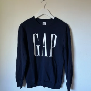 Mörkblå GAP sweatshirt med logga - Klassisk mörkblå sweatshirt från GAP med stor vit logga framtill. Tröjan har rund halsringning, långa ärmar och ribbade muddar. Tillverkad i mjuk bomulls- och polyestermix som är skön att bära. Perfekt för en avslappnad och stilren look. 3R10