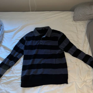 Randig pikétröja i blått och svart - Snygg långärmad pikétröja från Dressmann i marinblått och svart med breda ränder. Klassisk krage, knäppning vid halsen och en liten bröstficka. Perfekt för dig som gillar en sportig och avslappnad stil. Priset går att diskutera