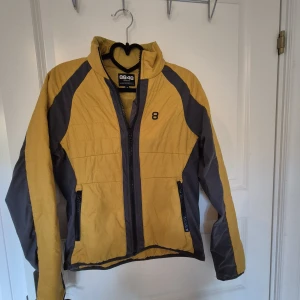Mellanlager  från 8848 Altitude - Sportig vindjacka från 8848 Altitude i gul och grå färg. Jackan har hel dragkedja framtill, två fickor med dragkedja och hög krage. Tillverkad i nylon och polyester för en lätt och smidig känsla. Perfekt för dig som gillar en aktiv stil. Stretchpaneler i sidorna och under ärmarna. 