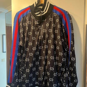 Svart bomberjacka från Gucci med GG-mönster - Säljer en svart bomberjacka från Gucci med vitt GG-mönster över hela jackan. Jackan har dragkedja framtill, ribbade muddar och krage med vita ränder samt röda och blå ränder längs ärmarna. Perfekt statement-plagg för dig som vill sticka ut.