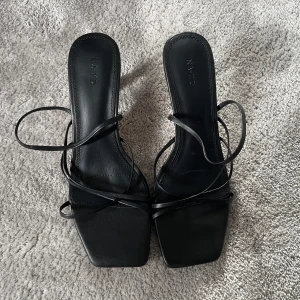 Svarta sandaletter med klack från NA-KD - Stilrena svarta sandaletter från NA-KD med smala remmar och fyrkantig tå. Skorna har en elegant låg klack och öppen tå, perfekt till sommarens outfits. Tillverkade i syntetmaterial med trendig design och snörning runt vristen. 7cm klack. Nypris 800kr, använda en gång.