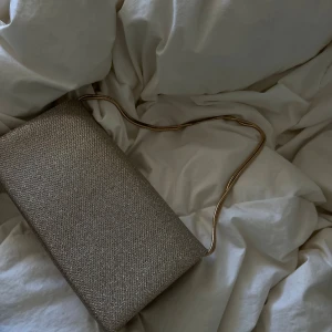 Glittrig guldig kuvertväska med kedja - Snygg kuvertväska i guld med glittrig yta och elegant guldfärgad kedja. Perfekt storlek för det viktigaste och ger en lyxig touch till din outfit. Stilren och enkel design som verkligen sticker ut.