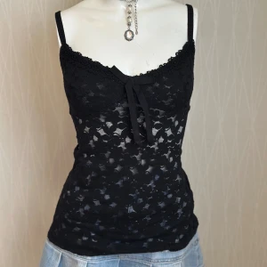 Svart spetslinne med rosett - Supersnygg svart linnetopp i spets med blommigt mönster och söt rosettdetalj framtill. Smala axelband och dekorativ kant upptill ger en feminin look. Perfekt att styla med jeans eller kjol för en trendig outfit.