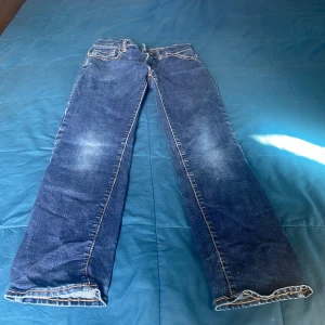 Blå raka jeans med kontrastsömmar - Säljer ett par klassiska blå jeans med raka ben och kontrastsömmar. Jeansen har fem fickor, bälteshällor och dragkedja med knapp. Materialet är robust denim och färgen har snygga slitningar på låren för en cool look.