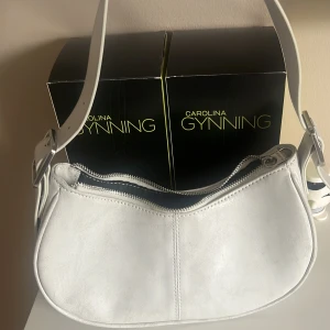 Vit axelväska i skinn med dragkedja - Snygg vit axelväska i skinn med bred justerbar axelrem och silvriga metalldetaljer. Väskan har en stilren, rundad form och stängs med dragkedja upptill. Perfekt för dig som vill ha en clean och modern look.