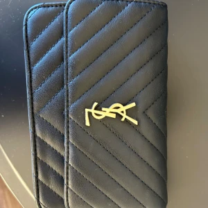 Svart YSL quiltad plånbok i skinn - Snygg svart quiltad plånbok från YSL med guldigt logotyp på framsidan. Tillverkad i äkta skinn med stilrent mönster och praktiskt fack på baksidan. Perfekt för dig som vill ha en lyxig och tidlös accessoar. Kommer med guldig kedja till!!