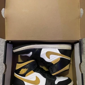 Nike Air Jordan 1 Mid svart/vit/guld - Säljer ett par Nike Air Jordan 1 Mid i storlek 41. Skorna har en snygg färgkombo i svart, vitt och guld med klassisk Jordan-logga på sidan. Ovandelen är i skinn och de har snörning framtill. Perfekta för dig som vill sticka ut med en ikonisk sneaker.
