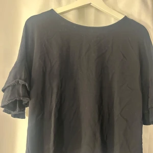 Svart blus med volangärmar och knappar - Svart blus i bomull med korta volangärmar och dekorativa guldiga knappar framtill. Blusen har en rund halsringning och en loose passform som ger en avslappnad vibe. Perfekt för dig som gillar stilrena plagg med en twist.