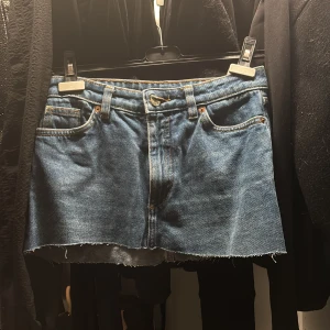 Blå jeanskjol med rå kant - Säljer en blå jeanskjol med klassisk femficksdesign och rå, avklippt kant nertill. Kjolen har bälteshällor, knapp och dragkedja framtill. Perfekt för dig som gillar en avslappnad och trendig denimlook.