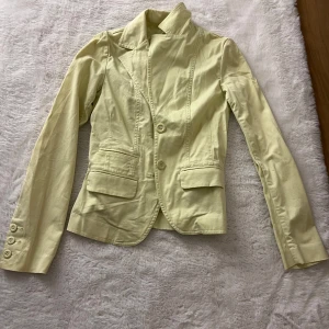 Ljusgul kostym med kavaj och byxor - Säljer en ljusgul kostym med figursydd kavaj och matchande utsvängda low waist byxor. Kavajen har två knappar, ficklock och klassisk krage. Byxorna har hög midja och vida ben. Perfekt för dig som vill sticka ut med färg! Materialet känns som bomull. Kavajen säljs separat för 299kr och båda tilsammans för 500kr