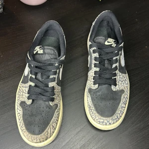 Nike Air Jordan 1 Low Elephant Print - Säljer ett par Nike Air Jordan 1 Low med ikoniskt elephant print i grått och svart. Skorna har svarta snören, vit swoosh och beige sula. Klassisk rund tå och lågt skaft, perfekt för dig som gillar streetwear och unika detaljer.