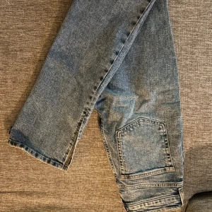 Blå jeans med slits från H&M, strl 34 - Säljer ett par blå jeans från H&M i storlek 34. Byxorna har en bredare passform med slits nertill på benen och en stor ficka på sidan. Perfekta för dig som gillar en avslappnad och trendig look.