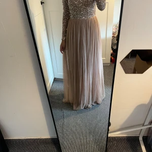 Två långklänningar med spets och glitter - Säljer två eleganta långklänningar. Ena är champagnefärgad med glittrig överdel och lång ärm, kjolen är mjukt plisserad. Den andra är svart med spetsdetaljer, v-ringning och lång ärm, samt flowy kjol. Perfekta för bal eller festliga tillfällen.