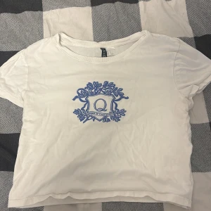Vit t-shirt med blå tryck från H&M Divided - Vit t-shirt från H&M Divided med blått tryck framtill som föreställer en sköld med bokstaven Q och texten 'Queen of your heart'. Klassisk rund halsringning och korta ärmar. Tillverkad i mjukt material, perfekt för en avslappnad stil.