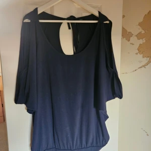 Mörkblå topp med öppen rygg från Rainbow - Säljer en mörkblå topp från Rainbow med lös passform, knytband i nacken Toppen har korta, vida ärmar öppet på överärmarna och bred resår nertill för en avslappnad look. Perfekt för dig som gillar stilrena och bekväma plagg.