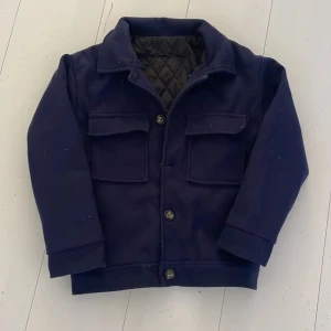 Mörkblå Fältjacka - Mörkblå jacka med krage och stora fickor framtill. Jackan har knäppning med knappar och quiltat innerfoder. Perfekt för kyligare dagar och har en klassisk, stilren look. För 12-13 år.