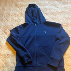 Marinblå ziphoodie från Polo Ralph Lauren - Snygg marinblå ziphoodie från Polo Ralph Lauren med dragkedja och klassisk huva. Tröjan har långa ärmar, fickor framtill och den ikoniska broderade loggan på bröstet. Perfekt för dig som gillar stilren och bekväm streetwear. Sitter löst, passar även som M!