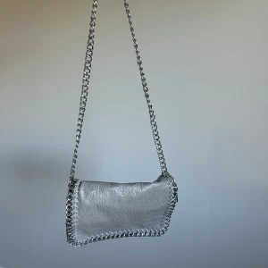 Grå handväska - Snygg axelväska i silverfärgat skinn från Tiamo. Väskan har en kedja som axelrem och kedjedetaljer längs kanterna. Magnetlås och innerfack med dragkedja. 