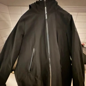 Svart vindjacka från Michael Kors - Svart vindjacka från Michael Kors med huva och snygga dragkedjor framtill. Jackan har en stilren look med diskret logga på bröstet och praktiska fickor med dragkedja. Perfekt för dig som gillar minimalistisk och urban stil.