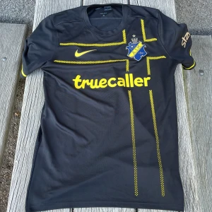 AIK fotbollströja Nike svart/gul M - AIK matchtröja från Nike i svart med gula detaljer och rutmönster. Snyggt AIK-emblem på bröstet, gul Nike-logga och sponsortryck. Tillverkad i lätt och ventilerande Dri-FIT-material, rund hals och korta ärmar. Perfekt för dig som vill representera AIK på läktaren eller träningen.