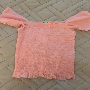 Rosa smockad croppad topp Cubus - Söt croppad topp från Cubus i ljusrosa med smockad struktur och volangkant nertill. Offshoulder-modell med korta, vida ärmar och liten rosett framtill. Perfekt för en somrig look och lätt att matcha med jeans eller kjol.