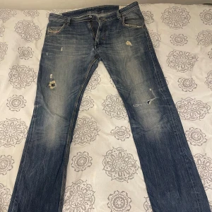 Blå Diesel Krooley jeans W33 L32 - Säljer ett par blå Diesel Krooley jeans med slitna detaljer och hål på benen. Jeansen har klassisk femficksdesign, orange kontrastsömmar och en snygg tvättad look. Modellen är rak och avslappnad med normal passform. Perfekta för dig som gillar en cool och avslappnad stil.