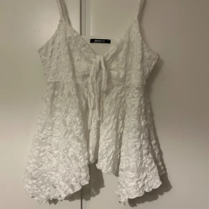 Vit spetsig linnetopp från Gina Tricot - Superfin vit linnetopp från Gina Tricot med spetsdetaljer och knyt framtill. Toppen har smala axelband och ett blommigt spetsmönster som ger en romantisk vibe. Perfekt för varma dagar och somriga outfits.