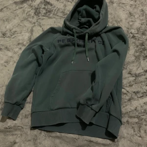 Mörkgrön hoodie med broderad text - Snygg mörkgrön hoodie från Peak Performance med broderad text på bröstet. Tröjan har huva med snörning och en stor magficka. Perfekt för chill dagar eller när du vill vara extra bekväm.