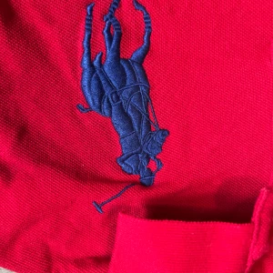 Röd pikétröja från Ralph Lauren - Klassisk röd pikétröja från Ralph Lauren med stor blå broderad logga på bröstet. Tröjan har krage, korta ärmar och knäppning med två knappar. Tillverkad i bomull och har en snygg, sportig look som sticker ut.