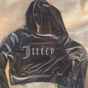 Juicy Couture svart velour hoodie - Svart velour hoodie från Juicy Couture med glittrande Juicy-tryck på ryggen. Jackan har huva, dragkedja framtill och två fickor. Mjuk och glansig finish, perfekt för en chill och trendig look.