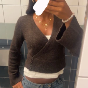 Brun omlottkofta med knappar - Mysig brun omlottkofta med v-ringning och fyra dekorativa knappar på sidan. Koftan är croppad och har långa ärmar med lite vidare avslut. Perfekt att styla med jeans eller kjol för en trendig look.