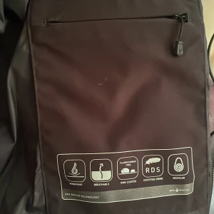 Svart dunjacka från Sail Racing - Svart pufferjacka från Sail Racing med huva och dragkedja. Jackan är fylld med certifierat dun från Allied Feather + Down (600 fill power) och har vind- och vattenavvisande yttertyg. Snygg logga på bröstet och bak på huvan. Perfekt för kalla vinterdagar.