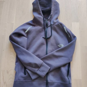 The North Face hoodie - Säljer en lila hoodie från The North Face med dragkedja och huva. Tröjan har långa ärmar, två fickor med dragkedja och reflekterande detaljer på axlarna. Perfekt för kyliga dagar och snygg streetstil.