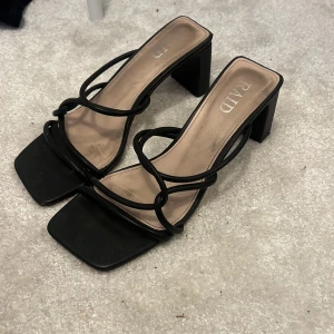 Svarta sandaler med klack från RAID - Säljer ett par svarta sandaler från RAID med fyrkantig tå och klack. Skorna har tunna, svarta remmar i snyggt mönster över foten och öppen tå. Snygg design som ger en trendig vibe och passar perfekt till sommarens outfits.