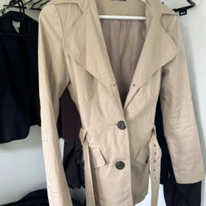Beige trenchcoat med bälte - Stilren beige trenchcoat med stora slag, knappar framtill och tillhörande bälte i midjan. Jackan har långa ärmar och fickor med lock, perfekt för dig som vill ha en klassisk look.