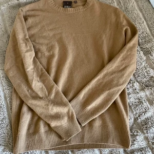 Beige stickad tröja från Oscar Jacobson - Säljer en klassisk beige stickad tröja från Oscar Jacobson, tillverkad i Italien. Tröjan har rund halsringning, ribbade muddar och är långärmad. Perfekt för lager-på-lager och enkel att matcha med jeans eller chinos.