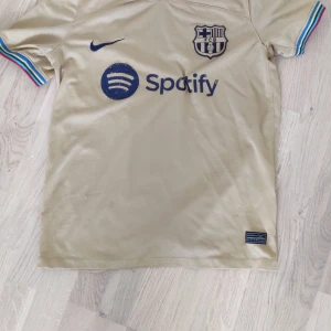 Beige FC Barcelona Nike fotbollströja - Snygg beige FC Barcelona fotbollströja från Nike med blå logga och Spotify-sponsor på bröstet. Ärmarna har färgglada ränder i blått, rött, gult och grönt. Materialet är lätt och andas, perfekt för match eller träning. UNHCR-tryck på ryggen.