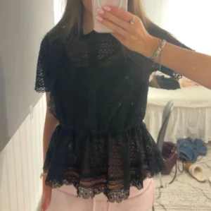 Svart spetsblus med volang - Säljer en svart blus i spets med korta ärmar och volang nertill. Blusen har fina spetsdetaljer och ett lätt transparent mönster som ger en romantisk vibe. Perfekt att styla med jeans eller kjol för en trendig look.
