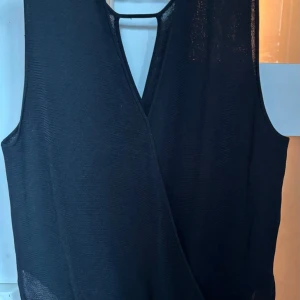 Svart omlottopp från Tiger of Sweden - Snygg svart omlottopp från Tiger of Sweden med v-ringning och ärmlös design. Toppen har en stilren och minimalistisk look med ett tunt, lätt material som ger en luftig känsla. Perfekt för dig som gillar klassiska och enkla plagg med en twist.