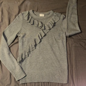 Grå stickad tröja - Grå stickad tröja från Vila i storlek M. Tröjan har långa ärmar och är dekorerad med en snygg volangdetalj som går diagonalt över framsidan och längs halsringningen. Fint skick utöver en tråd som börjat gå upp vid ena axeln (se bilder). Vill påstå att den är liten i storleken men passar mig fint som annars bör storleken xs-s 🤗