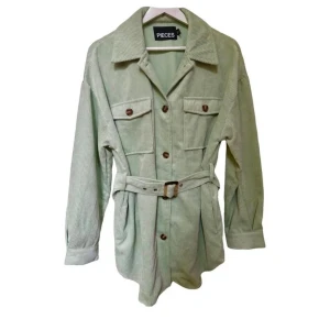 Grön manchester overshirt från Pieces - Snygg ljusgrön overshirt från Pieces i manchester med bälte i midjan och stora bröstfickor. Jackan har knappar framtill, klassisk krage och långa ärmar. Perfekt för lager-på-lager och ger en chill men trendig vibe.