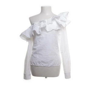 Vit offshoulder blus med volang - Snygg vit blus med offshoulder-design och stora volanger längs halsringningen. Toppen har långa ärmar och en stilren, feminin look som passar perfekt till jeans eller kjol. Tillverkad i bomull för en fräsch känsla.