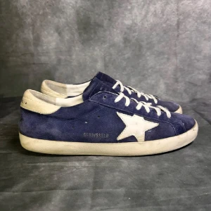 Blå golden goose skor - Snygga blå sneakers från Golden Goose med vit stjärndetalj på sidan och vita snören. Skorna har en klassisk låg profil och är gjorda i mocka med vit sula och häl. Perfekta för dig som gillar streetstyle och vill sticka ut lite extra.