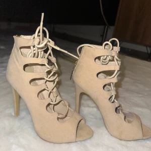 Beige klackskor från Public Desire - Snygga beige klackskor från Public Desire med höga smala klackar, snörning framtill och öppen tå. Skorna har mockaliknande material och dragkedja bak för enkel påtagning. Perfekta för dig som vill sticka ut med trendiga detaljer.