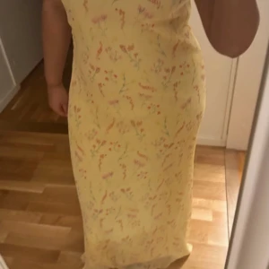 Gul blommig långklänning från NA-KD - Superfin långklänning från NA-KD i ljusgul nyans med blommigt mönster. Klänningen har en rak passform och är ärmlös, perfekt för varma dagar. Materialet känns lätt och luftigt, vilket gör den skön att bära hela sommaren.