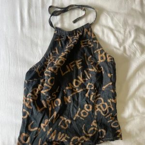 Svart halterneck topp med textmönster - Cool svart topp med halterneck och bruna texttryck över hela tyget. Toppen har dragkedja i ryggen och knytning i nacken. Snygg, edgy look som sticker ut och passar perfekt till jeans eller kjol.