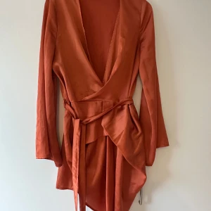 Rostfärgad omlottklänning med bälte - Snygg rostfärgad omlottklänning från Missguided i satinliknande material. Klänningen har långa ärmar, djup v-ringning och knytband i midjan som ger en smickrande siluett. Perfekt för dig som vill sticka ut med en trendig och elegant look.