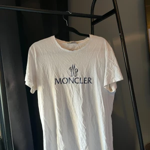 Vit Moncler t-shirt med logga - Vit t-shirt från Moncler i bomull med klassisk rund hals och stor mörkblå Moncler-logga framtill. Enkel och stilren design, perfekt för dig som gillar exklusiva märken och clean look.
