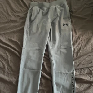 Grå träningsbyxor från Under Armour - Snygga grå träningsbyxor från Under Armour med logga på låret och text längs benet. Byxorna har resår i midjan, snörning och praktiska fickor. Perfekta för gymmet eller chill hemma, tillverkade i mjukt och stretchigt syntetmaterial.